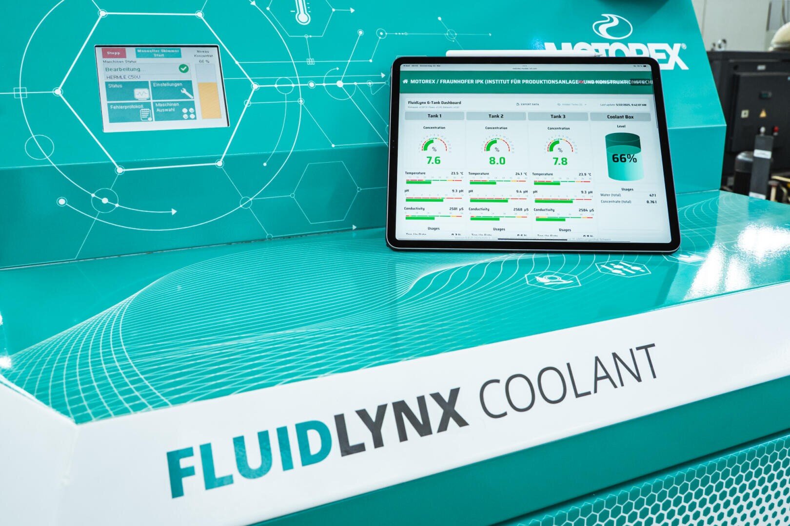 A tablet with performance displays stands on a blue-green machine labeled "FLUIDLYNX COOLANT", while control panels and digital interfaces can be seen in the background.