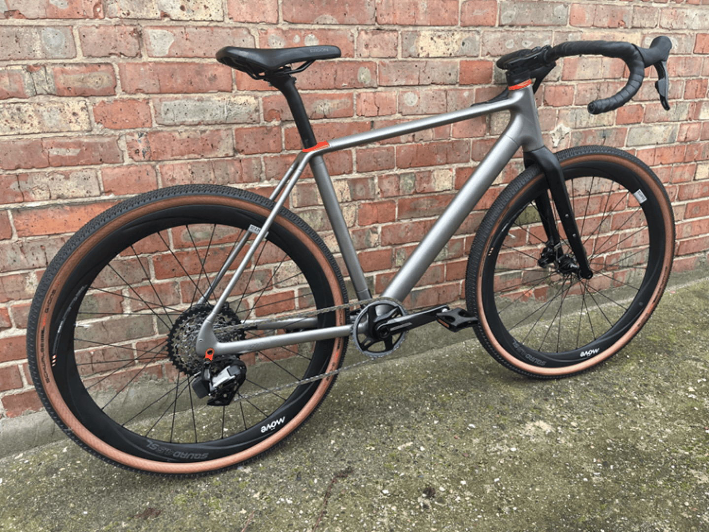 A modern silver and black road bicycle with tan sidewall tires is propped against a brick wall on a concrete surface. The bike features drop handlebars and a lightweight frame.