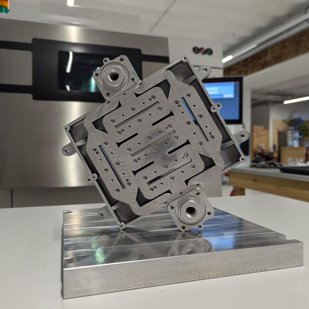 Close-up of a machined metal part with intricate grooves and patterns, mounted on a metal base. A modern industrial workspace with machines and screens can be seen in the background.