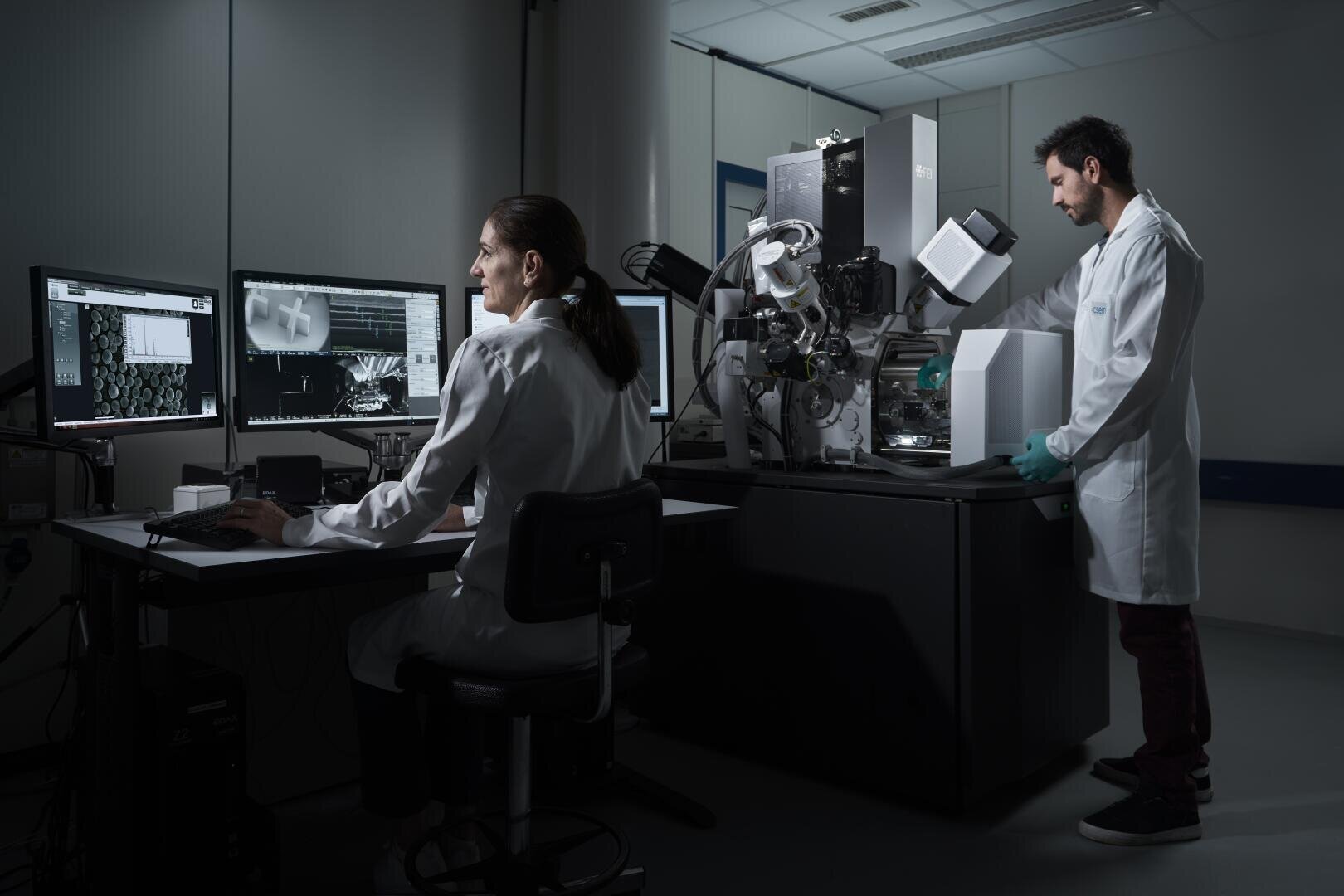 Two scientists in lab coats work in a dimly lit laboratory; one analyzes data on computer monitors while the other operates a large, advanced microscope or imaging machine.