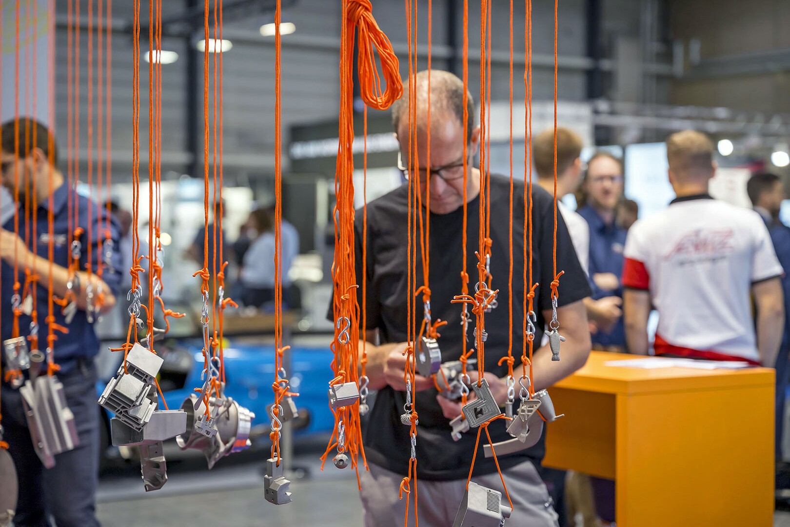 A man stands among numerous orange ropes hanging from above, each holding various metal objects. Other people and a yellow table are visible in the background at what appears to be an indoor event or exhibition.