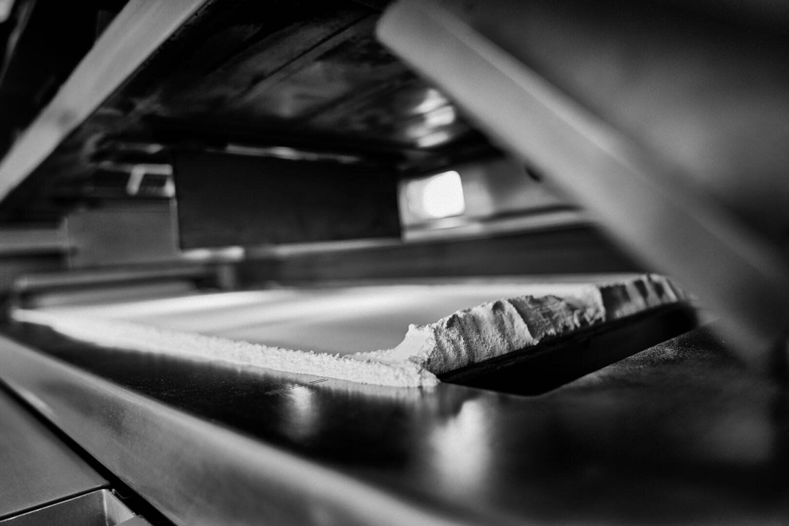 A close-up black and white photograph of a partially sliced loaf of bread on a metal surface under an industrial bread slicing machine.