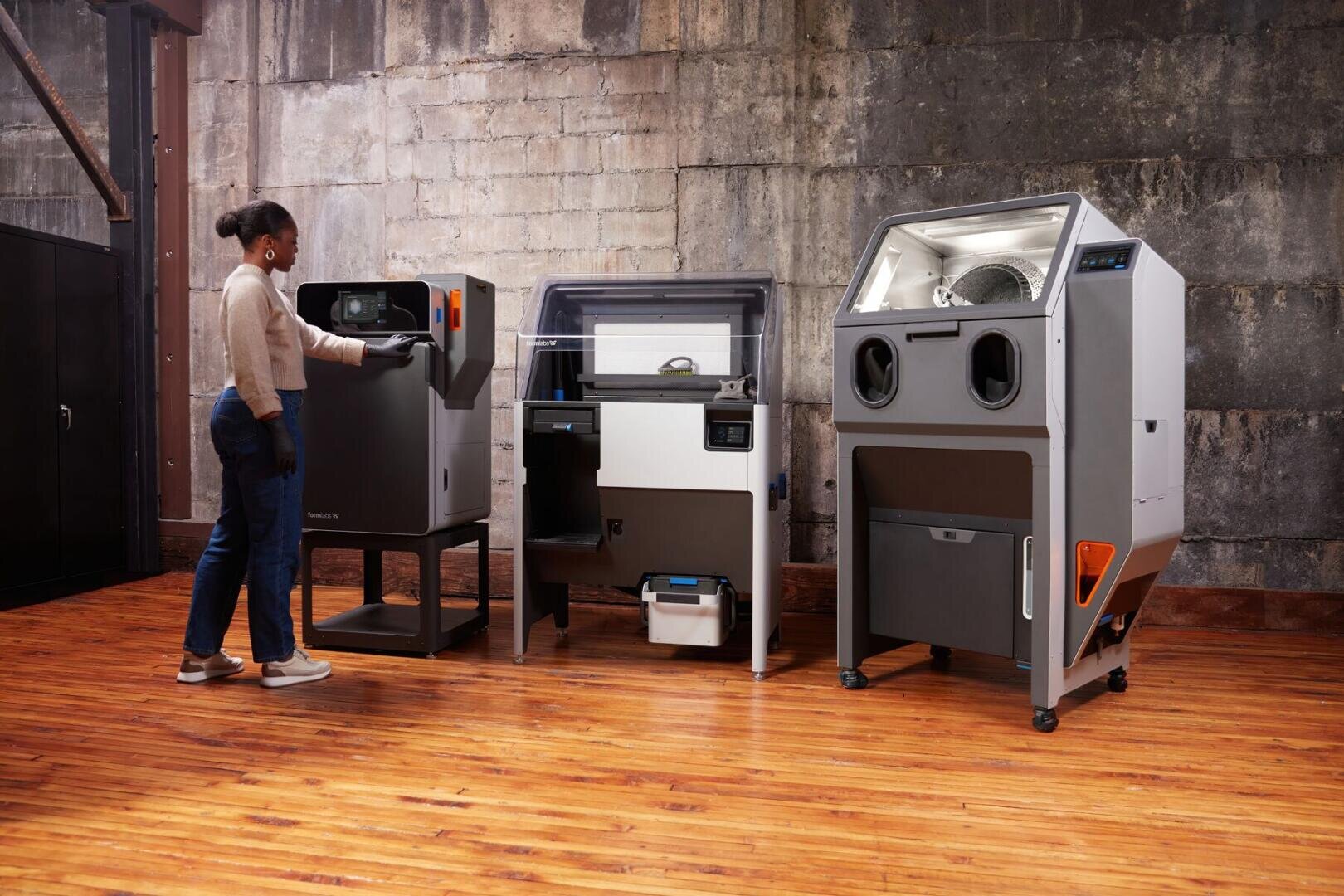 A woman operates a 3D printing machine in an industrial space with a wooden floor and concrete wall, next to two other large, modern machines.