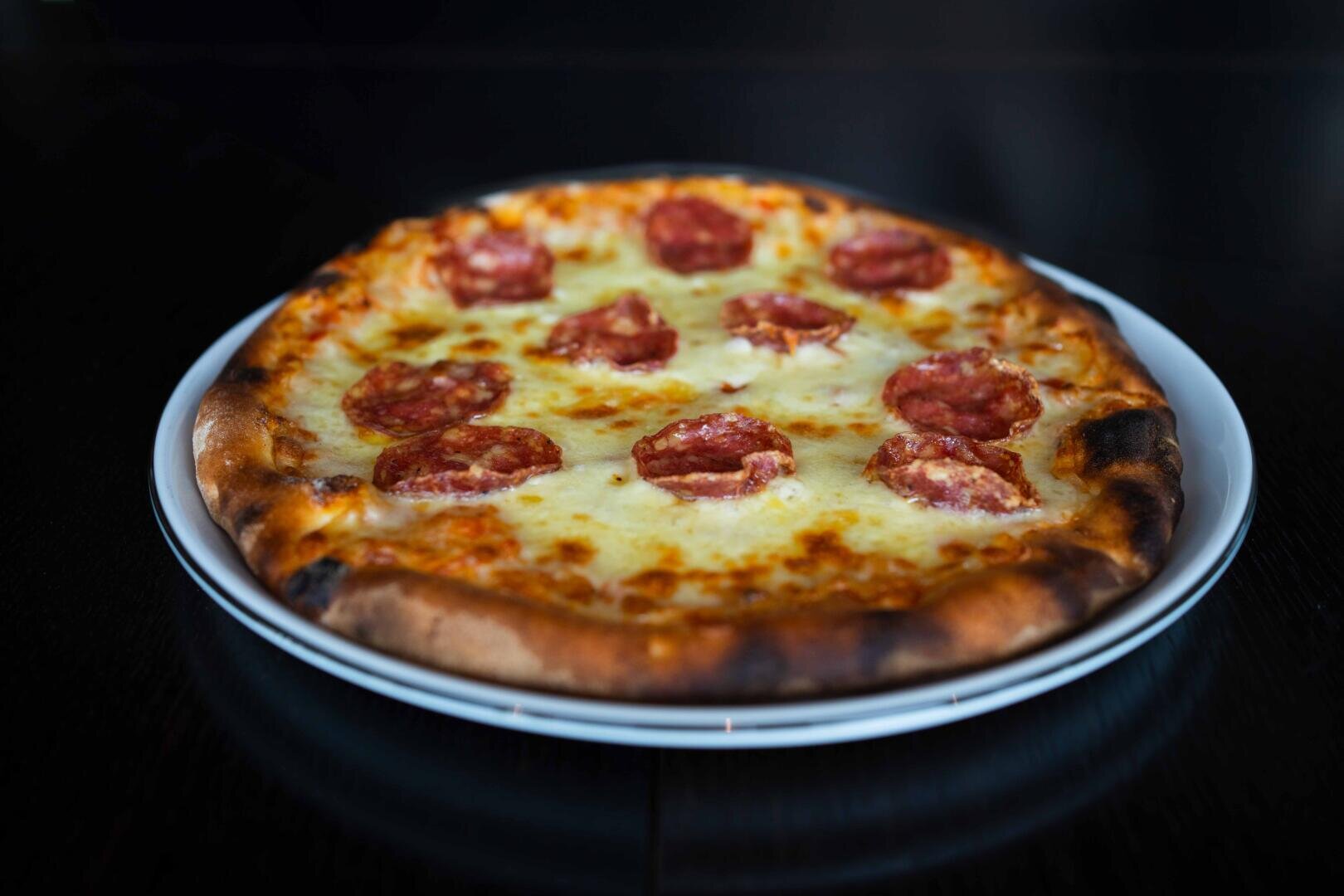 Eine Peperoni-Pizza mit geschmolzenem Käse und knusprigem Rand liegt auf einem weißen Teller vor einem dunklen Hintergrund.