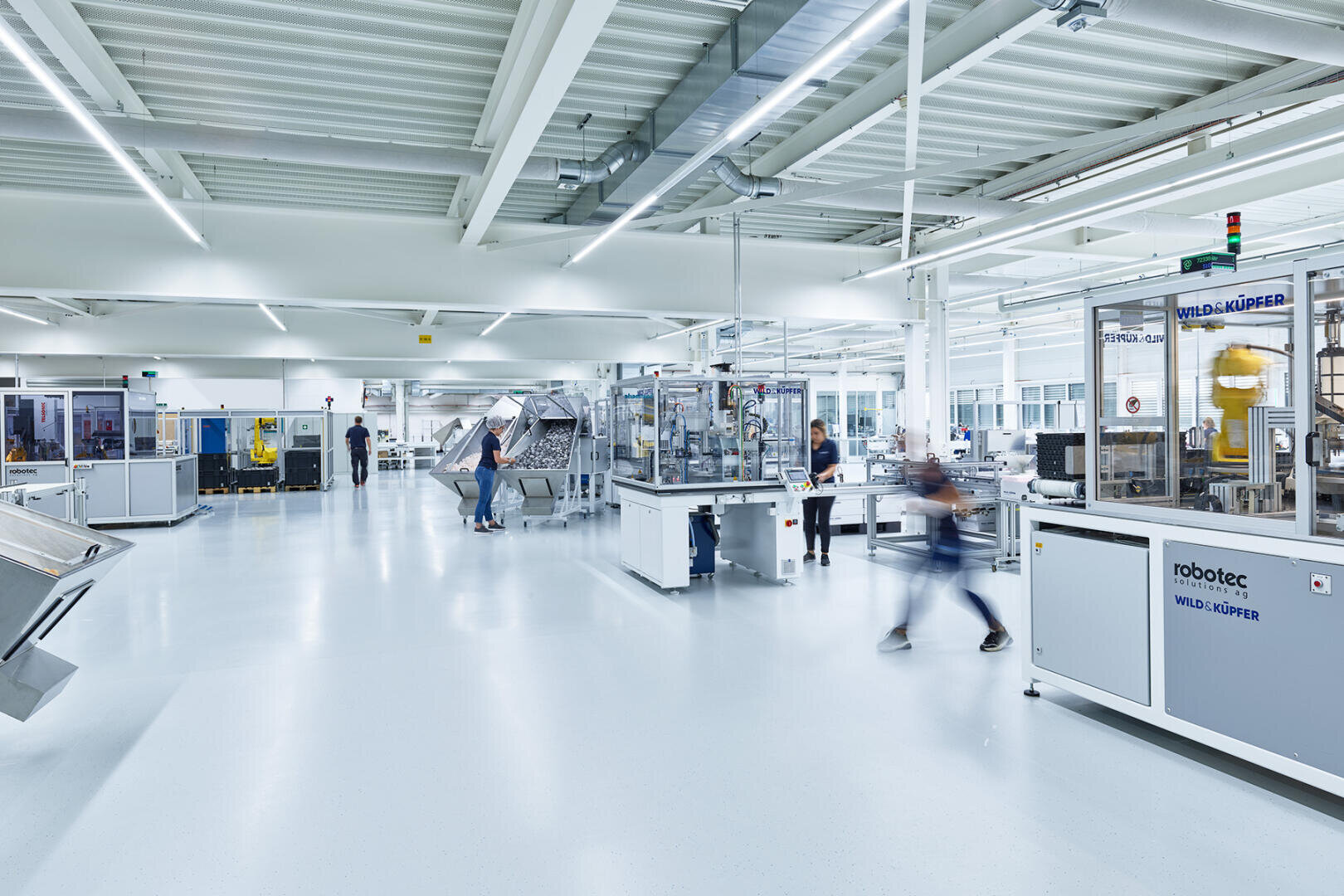 A bright, modern factory floor where people work among large industrial machinery and equipment, including robotic systems. The space is clean, spacious and well-lit with white floors and ceilings.