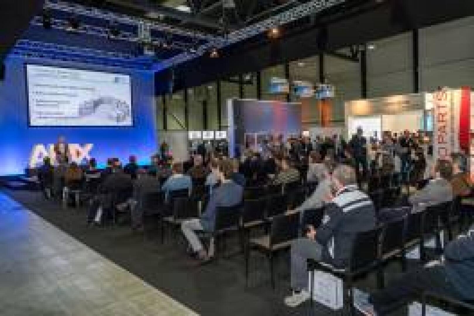 A large group of people sit in rows in front of a stage with a blue background and a projected presentation at a conference or seminar in a spacious, well-lit hall.