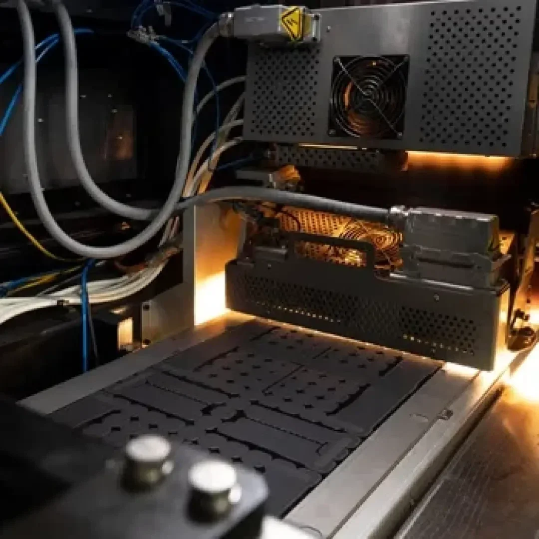 Nahaufnahme eines 3D-Druckers in Betrieb. Zu sehen sind beleuchtete Teile, Kabel und ein beheiztes Bett, auf dem Objekte gedruckt werden, in einer industriellen Umgebung.