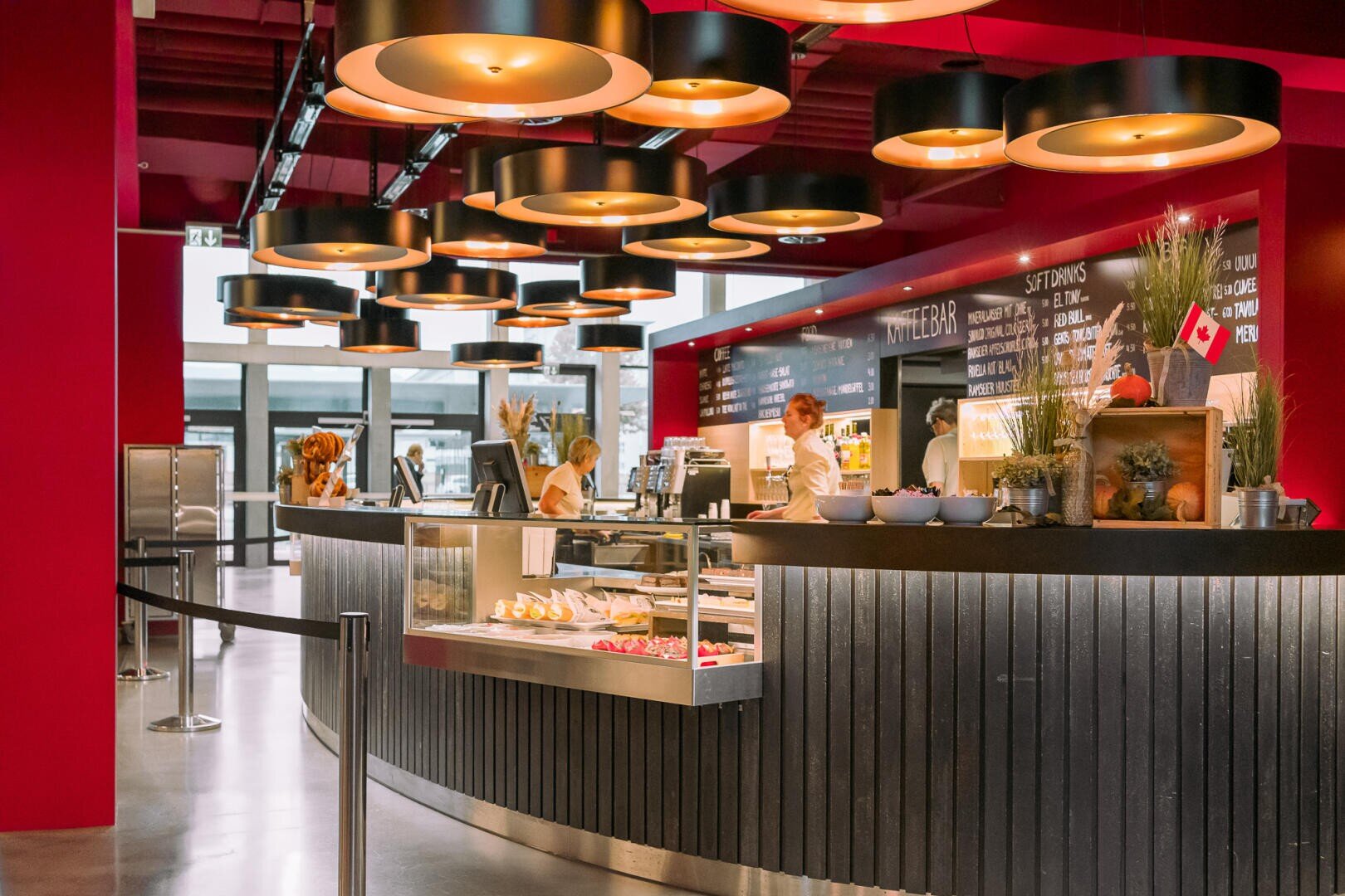 Ein modernes, hell beleuchtetes Café mit roten Wänden und mehreren runden Hängelampen. Das Personal bereitet die Speisen hinter einer geschwungenen Theke mit Gebäck zu, im Hintergrund stehen Menütafeln und Topfpflanzen.