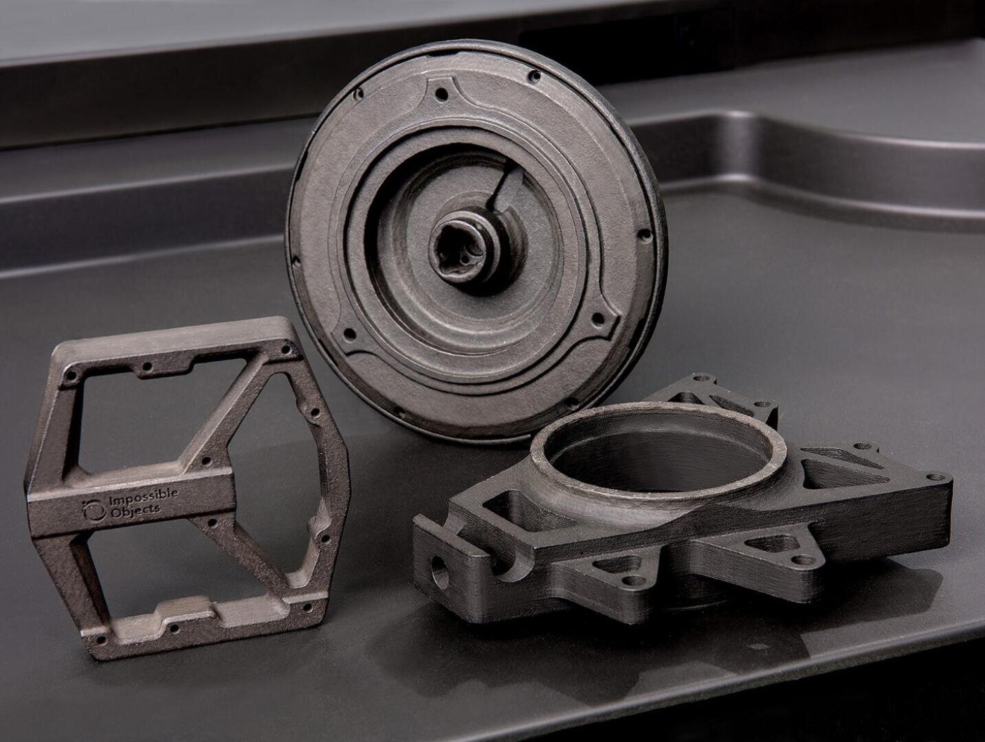 Drei schwarze, 3D-gedruckte mechanische Komponenten mit geometrischen Formen und komplizierten Designs werden auf einer mattschwarzen Oberfläche ausgestellt. Die Objekte wirken haltbar und haben eine leicht strukturierte Oberfläche.