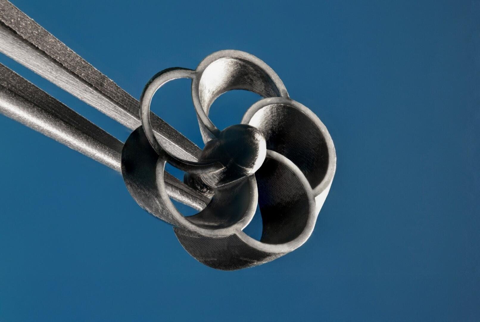Close-up of a metallic object with curved, interlocking loops and rods against a solid blue background, resembling an abstract sculpture or a piece of modern art.