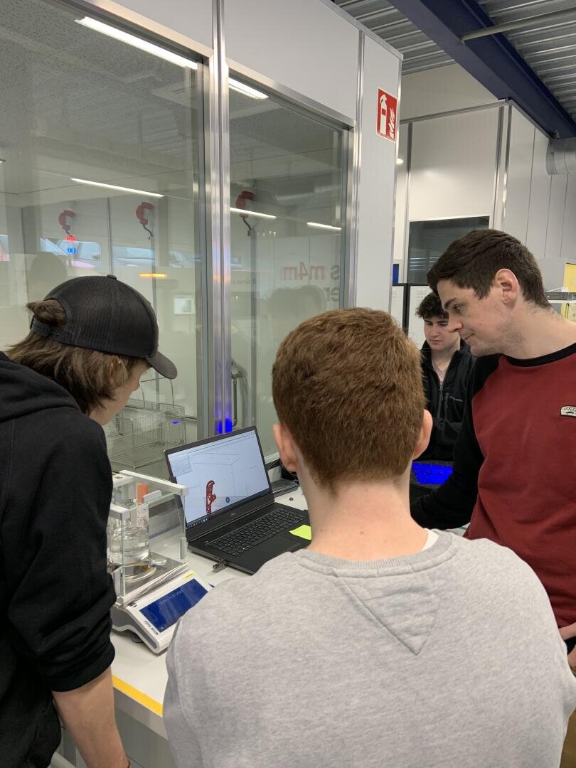 Four people stand around a table, looking at a laptop displaying a 3D model. One person is holding a phone, and laboratory equipment is visible on the table in a modern, glass-walled workspace.