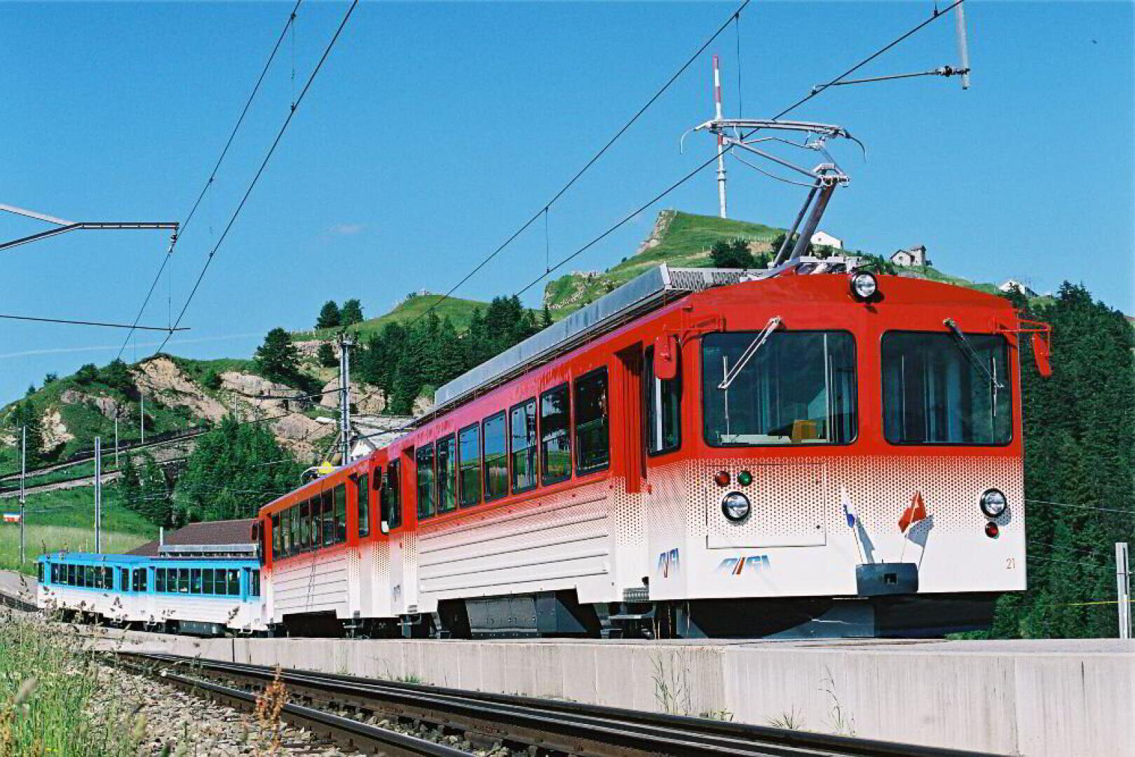 Ein rot-weißer elektrischer Zug, der auf Gleisen durch eine Berglandschaft fährt.