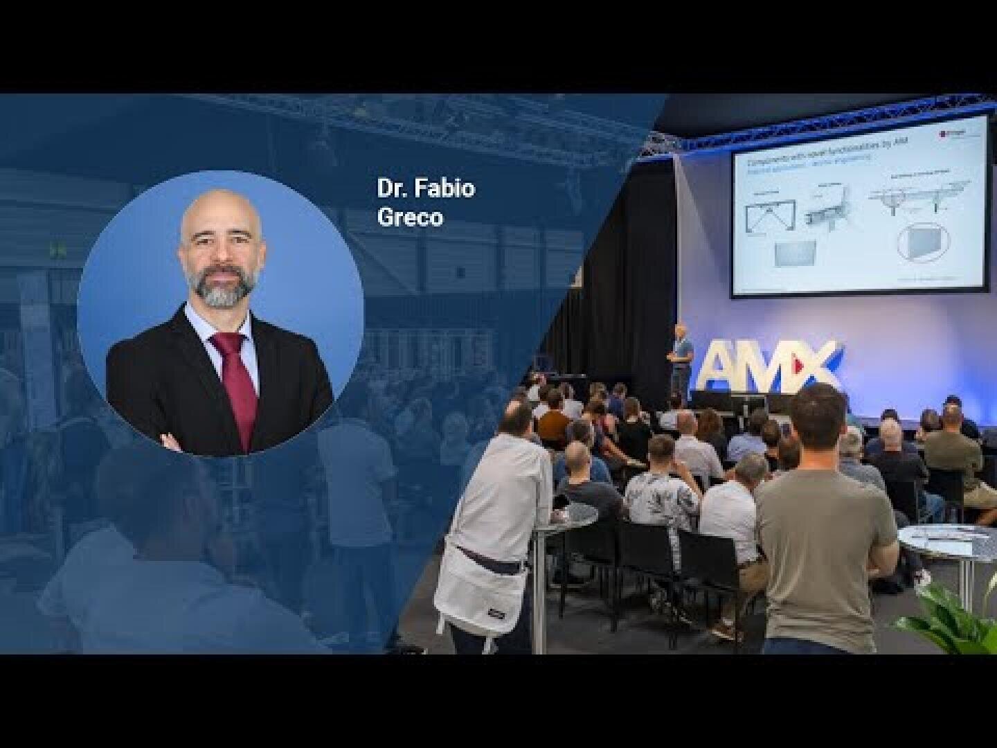 Ein großes Publikum verfolgt einen Vortrag auf einer Konferenz. Auf der linken Seite zeigt eine kreisförmige Grafik ein Porträt von Dr. Fabio Greco mit seinem Namen. Auf der Bühne präsentiert ein Redner Folien vor einem AMX-Schild.