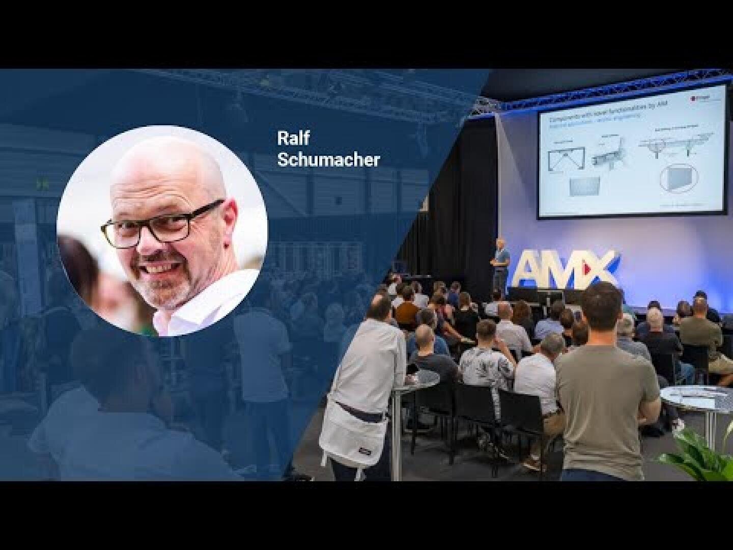 A speaker addresses the audience on stage at an event on which a screen with technical diagrams can be seen. A fade-in shows a headshot of a smiling man with a bald head and glasses, labeled Ralf Schumacher.