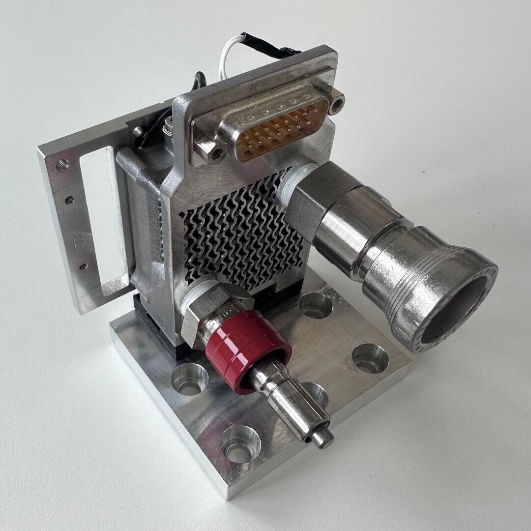 An industrial metal piece with a connecting piece at the top, a net-like center, red and silver fittings and a square base with four holes. It rests on a white surface.