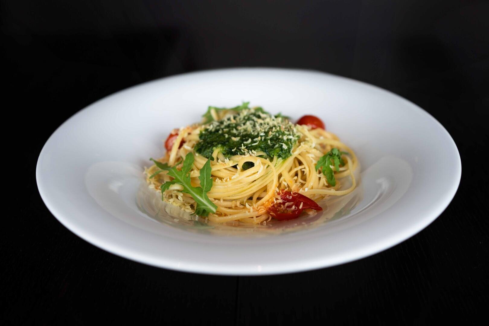 Ein weißer Teller mit Spaghetti, die mit grünem Pesto, geriebenem Käse, Kirschtomaten und Rucola belegt sind, vor einem dunklen Hintergrund.