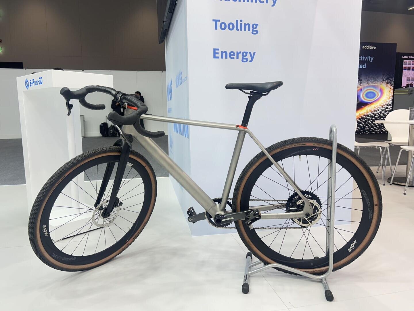 A modern silver road bike with drop handlebars and brown-edged tires is displayed on a metal stand indoors, next to a white wall with blue text that reads Machinery, Tooling, and Energy.