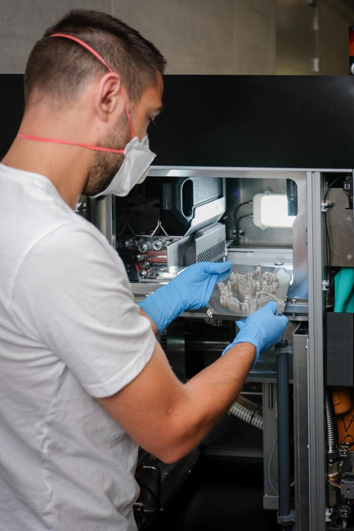 Eine Person, die eine Maske und blaue Handschuhe trägt, bedient einen 3D-Drucker und entnimmt vorsichtig ein transparentes Objekt aus dem Gerät in einem Labor oder einer industriellen Umgebung.