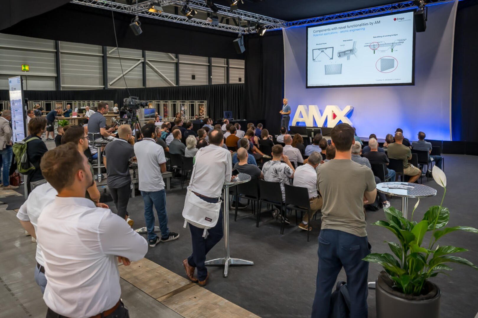 Eine große Gruppe von Menschen sitzt und steht in einem Veranstaltungssaal und beobachtet aufmerksam einen Redner, der auf der Bühne einen Vortrag hält, dessen Folien auf eine mit AMX beschriftete Leinwand projiziert werden.