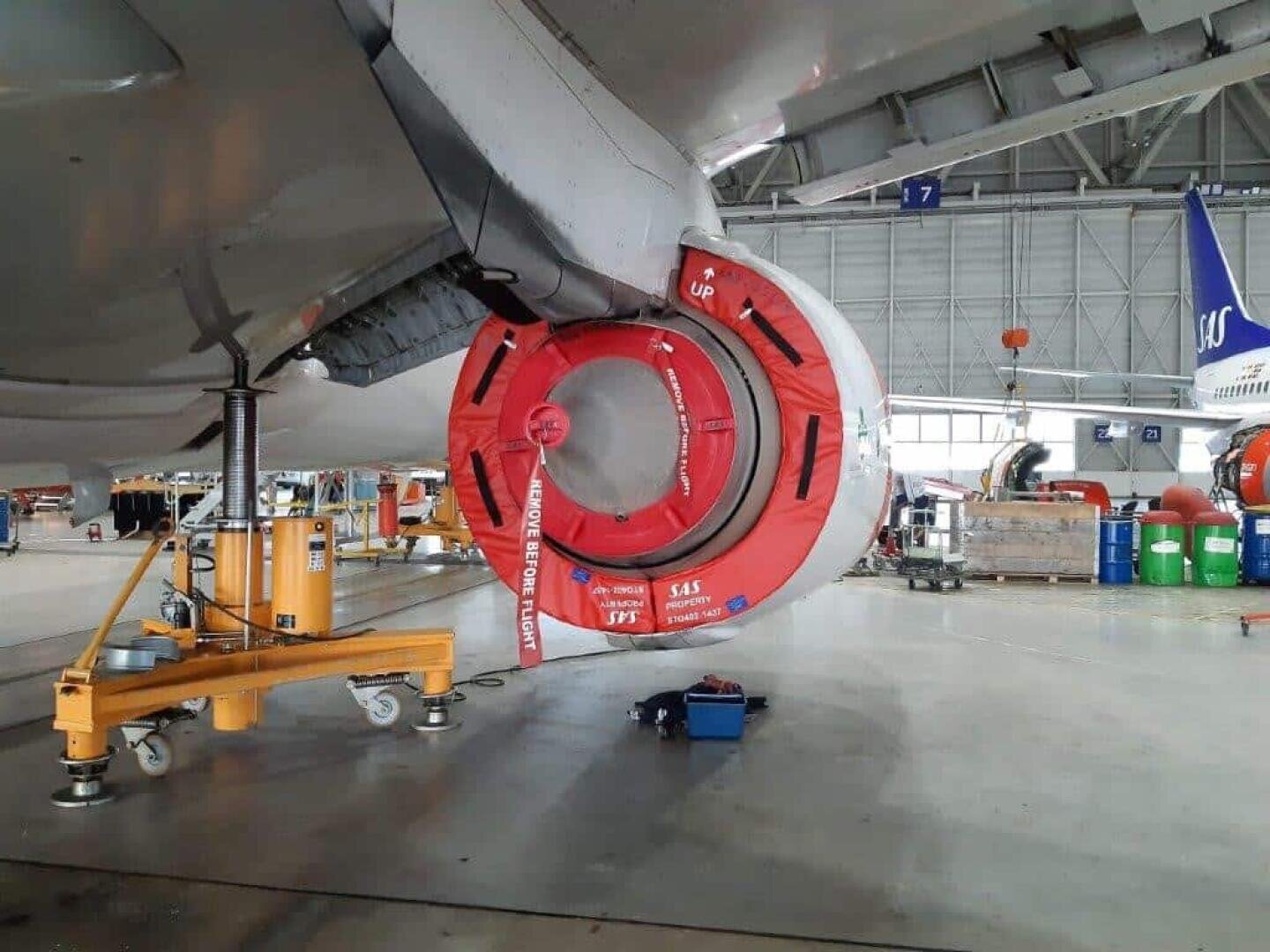 Nahaufnahme eines Triebwerks eines Verkehrsflugzeugs in einem Hangar. Über dem Triebwerkseinlass ist eine rote Schutzabdeckung mit der Aufschrift "Remove Before Flight" angebracht. Um den Bereich herum sind verschiedene Wartungsgeräte und Wagen zu sehen.