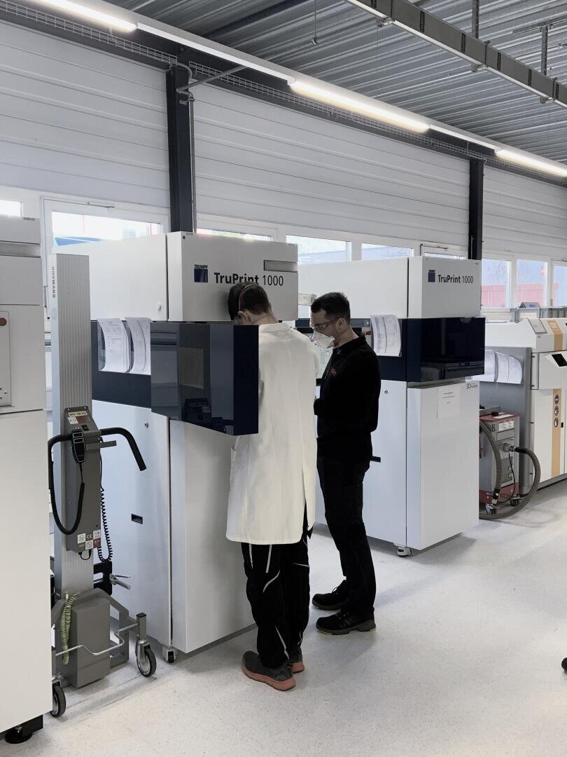Zwei Personen, eine im weißen Laborkittel und eine in dunkler Kleidung, stehen vor großen Industriemaschinen mit der Aufschrift TruPrint 1000 in einem modernen, gut beleuchteten Labor oder einer Produktionsstätte.