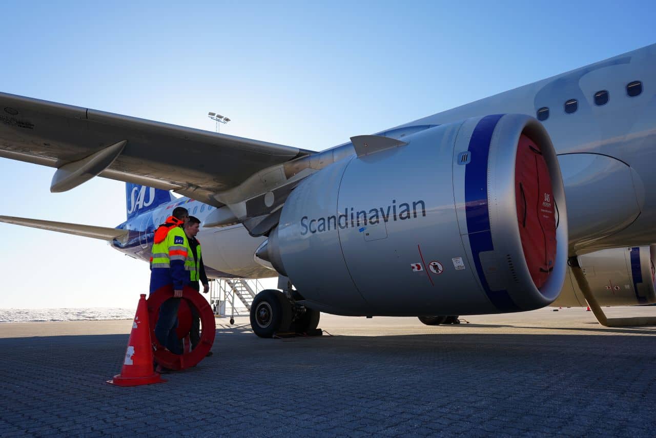 Ein Mitarbeiter des Bodenpersonals in einer reflektierenden Jacke steht in der Nähe eines SAS-Flugzeugtriebwerks, das abgedeckt auf einer sonnigen Flughafenrollbahn abgestellt ist. Neben dem Mitarbeiter sind orangefarbene Kegel auf dem Boden aufgestellt.