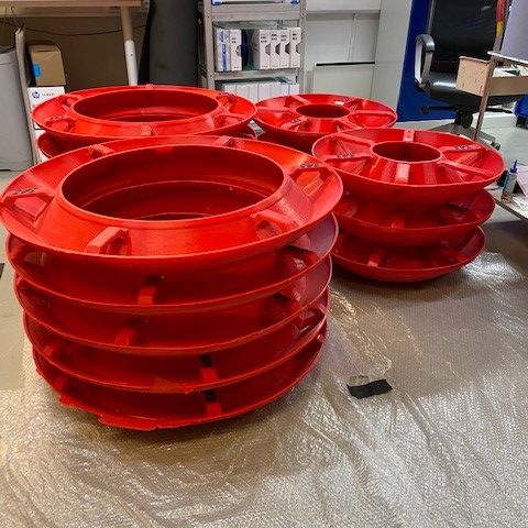 Vier Stapel großer, runder, leuchtend roter Plastikrollen liegen auf einer grauen Fläche in einem Arbeitsraum mit Regalen und Büromöbeln im Hintergrund.