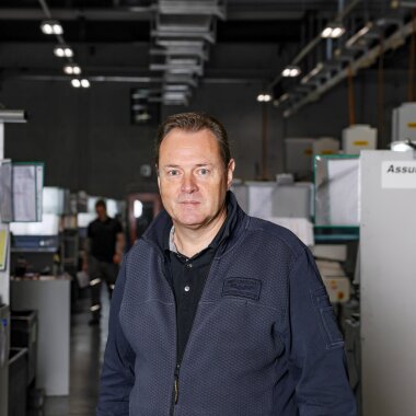 A man in a navy blue jacket stands in a well-lit industrial or factory environment with machinery and equipment in the background. The environment looks organized and professional.