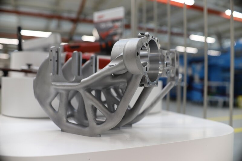 Ein komplexes, metallisches 3D-gedrucktes Teil mit geometrischen, gitterartigen Strukturen wird auf einer weißen Fläche in einer Industrie- oder Fertigungsumgebung dargestellt.