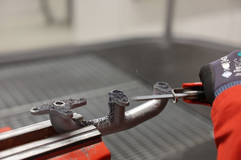 Eine Person, die einen Handschuh trägt, feilt oder entgratet mit einem Handwerkzeug die Kante eines in einem Schraubstock befestigten Metallteils, wobei auf der Oberfläche Metallspäne sichtbar sind.