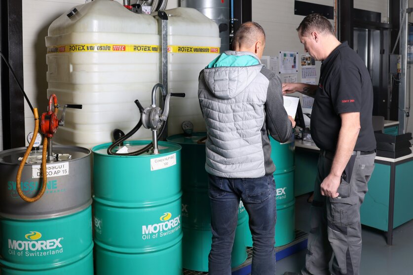Two men stand next to large Motorex oil barrels and storage tanks in a workshop; one man points at a clipboard while the other listens, with equipment and charts visible in the background.