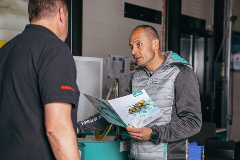 Zwei Männer unterhalten sich in einem Innenraum; einer von ihnen hält eine Broschüre mit dem Titel MOTIONTEX in der Hand, auf deren Umschlag Industrieteile abgebildet sind. Die Umgebung scheint ein Büro oder eine Werkstatt zu sein.