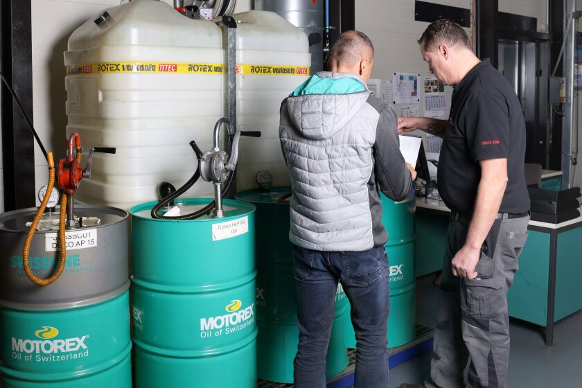 Zwei Männer stehen vor großen Öllagertanks und grünen Motorex-Ölfässern und besprechen in einer Industrieumgebung Papierkram. Einer trägt eine graue Weste, der andere ein schwarzes Hemd und eine graue Hose.
