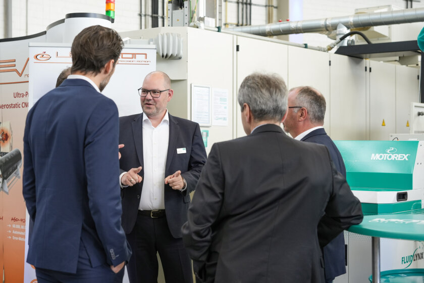 Five men in business suits stand in a group and discuss in an industrial or production environment in which machines and systems can be seen in the background.