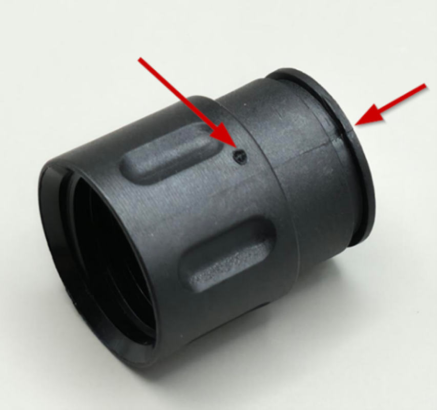 Ein schwarzer zylindrischer Kunststoffstecker mit strukturierten Griffbereichen ist auf weißem Hintergrund abgebildet. Zwei rote Pfeile weisen auf kleine Einkerbungen und Merkmale an der Seite und am Rand hin.