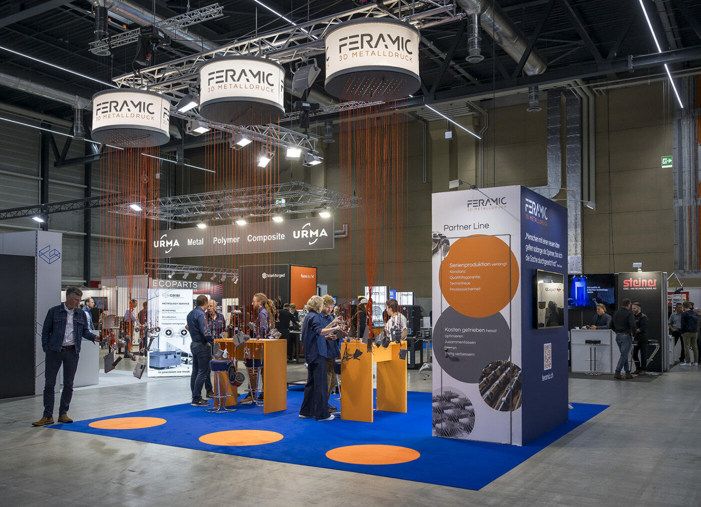 An einem Messestand von FERAMIC zum Thema 3D-Metalldruck werden Produktmuster und Informationen präsentiert. In einer modernen Messehalle können Besucher unter markenspezifischer Beleuchtung und Beschilderung mit den Mitarbeitern interagieren.