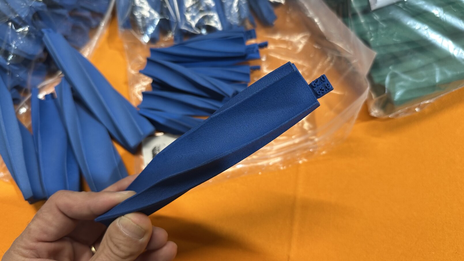 Eine Hand hält einen flachen, nicht aufgeblasenen blauen Luftballon über einem Tisch, der mit weiteren blauen und grünen, nicht aufgeblasenen Luftballons in Plastiktüten bedeckt ist.