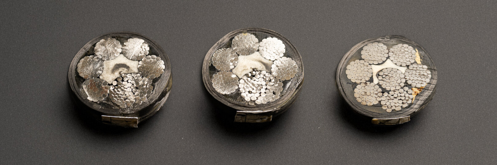 Drei Querschnitte dicker elektrischer Kabel sind nebeneinander auf einer dunklen Fläche abgebildet und zeigen dicht gepackte, silberfarbene Metalldrähte, die in kreisförmigen Mustern in jedem Kabel angeordnet sind.