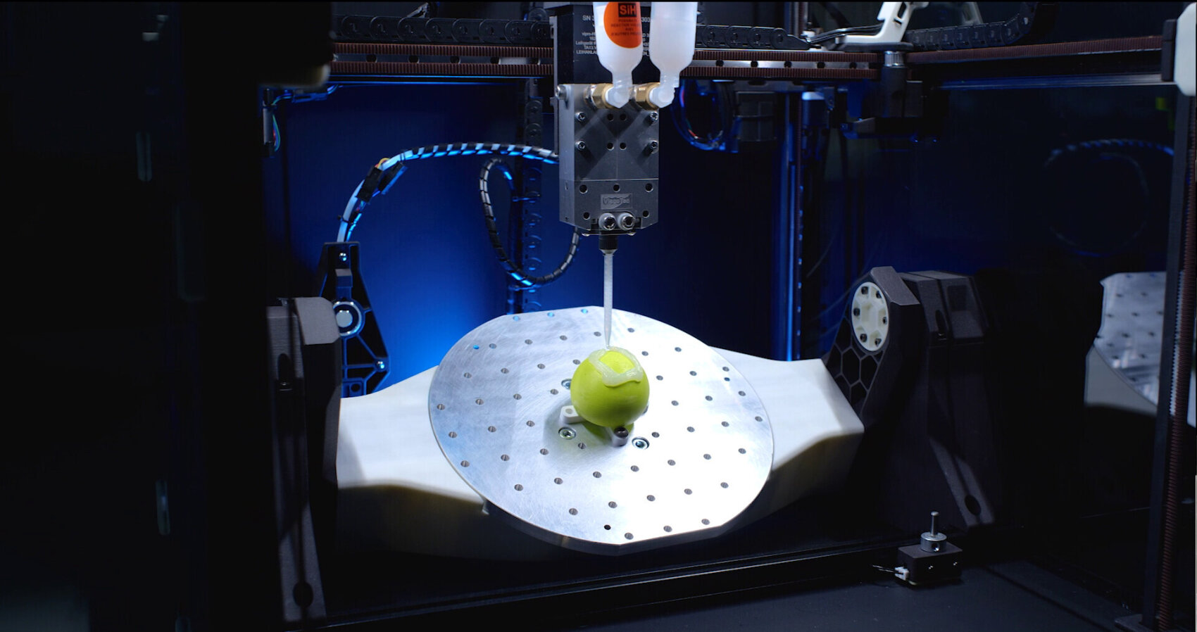 Ein 3D-Drucker extrudiert Material auf einen grünen Apfel, der auf einer rotierenden Metallplattform steht, während blaues Licht die Maschine und die umliegenden Komponenten beleuchtet.