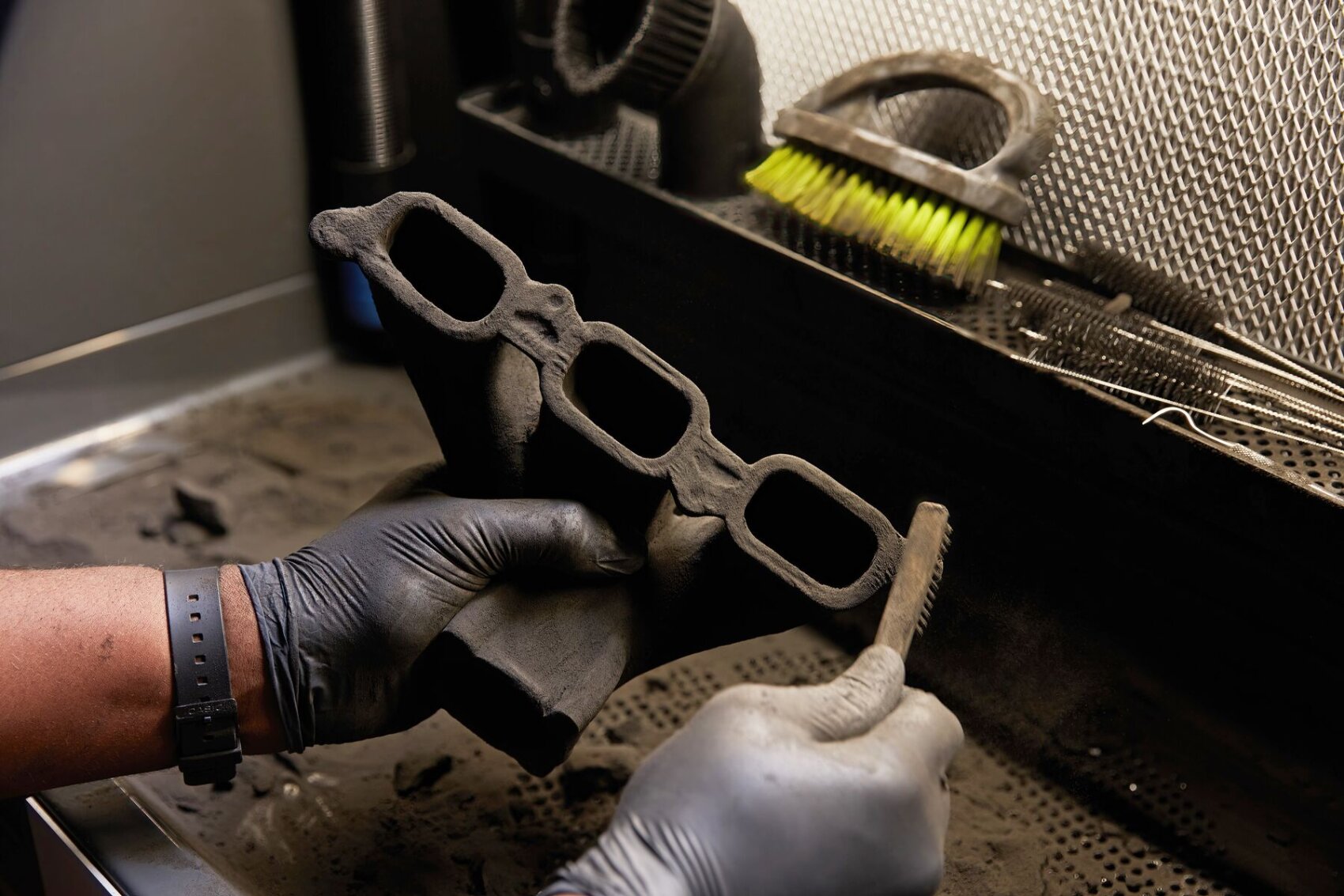 A person wearing black gloves cleans a dusty, 3D-printed car part in a workshop with a brush, while other cleaning tools can be seen in the background.