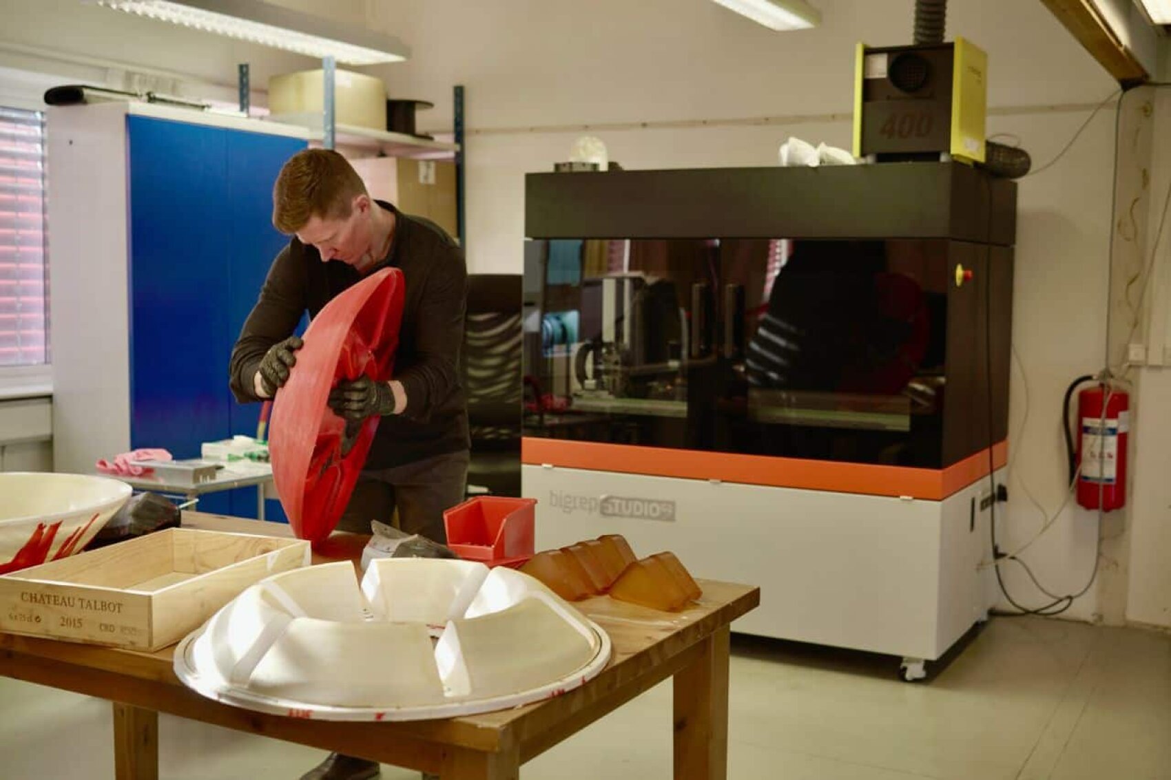 Eine Person mit Handschuhen inspiziert ein großes rotes 3D-gedrucktes Objekt in einer Werkstatt mit einem 3D-Drucker, Arbeitstischen, Kisten und einem weiteren weißen Objekt mit ähnlicher Form auf dem Tisch.