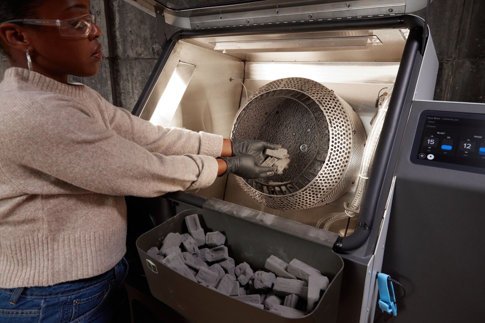 A person wearing gloves places or removes dark cylindrical objects in a large metal machine, while other similar objects are collected in a container below. A control panel with a display can be seen on the right-hand side.